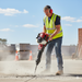 A construction worker in a high-visibility vest and ear protection uses the Paddock Motorised Jackhammer by Paddock to break concrete outdoors, sending dust and debris flying on a sunny day.