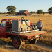A man in a plaid shirt and straw hat stands in a dry field beside a red flatbed truck carrying a Paddock Fire Fighting Tank by Paddock. A barn and trees are seen in the background under a clear sky.
