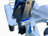 A Paddock Mini Loader & Excavator Alloy Equipment Trailer, featuring a diamond plate surface and white wheel fenders, carries a hydraulic attachment secured with blue and yellow straps to its front.