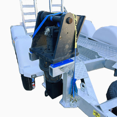 A Paddock Mini Loader & Excavator Alloy Equipment Trailer, featuring a diamond plate surface and white wheel fenders, carries a hydraulic attachment secured with blue and yellow straps to its front.