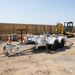 A Paddock Mini Loader & Excavator Alloy Equipment Trailer is parked on a gravel lot at a construction site, surrounded by orange cones, wooden planks, tools, and a yellow mini excavator under a clear sky.