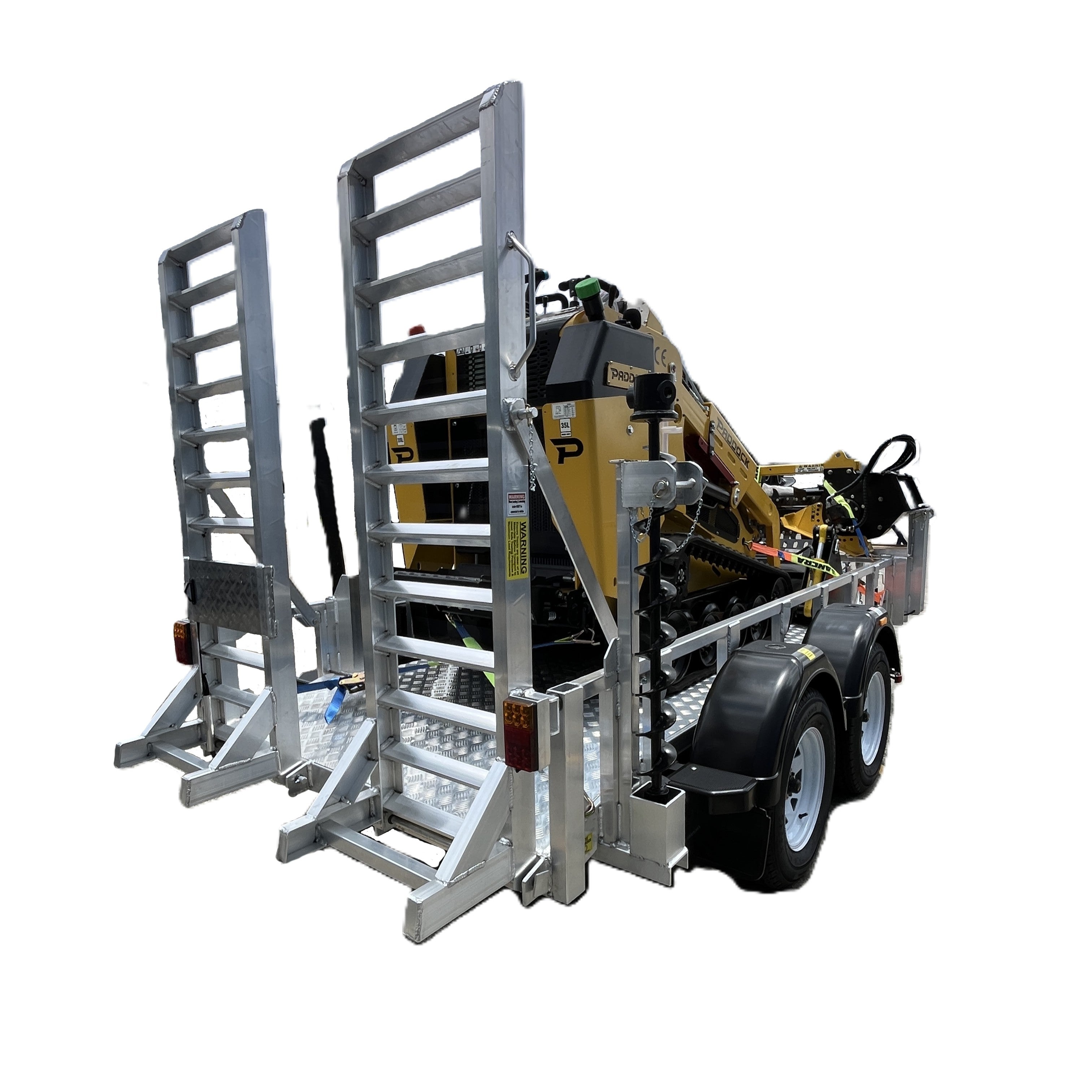A Paddock Mini Loader & Excavator Alloy Equipment Trailer by Paddock, featuring dual ramps, transports yellow construction gear. Parked on concrete near an industrial building, it's built to handle tough jobs.