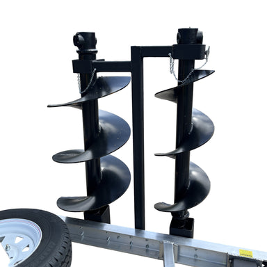 Two large black auger drill bits are mounted vertically on a metal frame from the Paddock Mini Loader & Excavator Alloy Equipment Trailers by Paddock. Part of a custom alloy trailer and a white tire are visible at the bottom left against a white background.