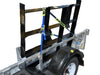 A Paddock Mini Loader & Excavator Alloy Equipment Trailer by Paddock, Spreader bar with black metal supports, blue and yellow straps, diamond plate flooring, and a white wheel, is parked on concrete near a blue wall.