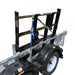 A Paddock Mini Loader & Excavator Alloy Equipment Trailer by Paddock, Spreader bar with black metal supports, blue and yellow straps, diamond plate flooring, and a white wheel, is parked on concrete near a blue wall.