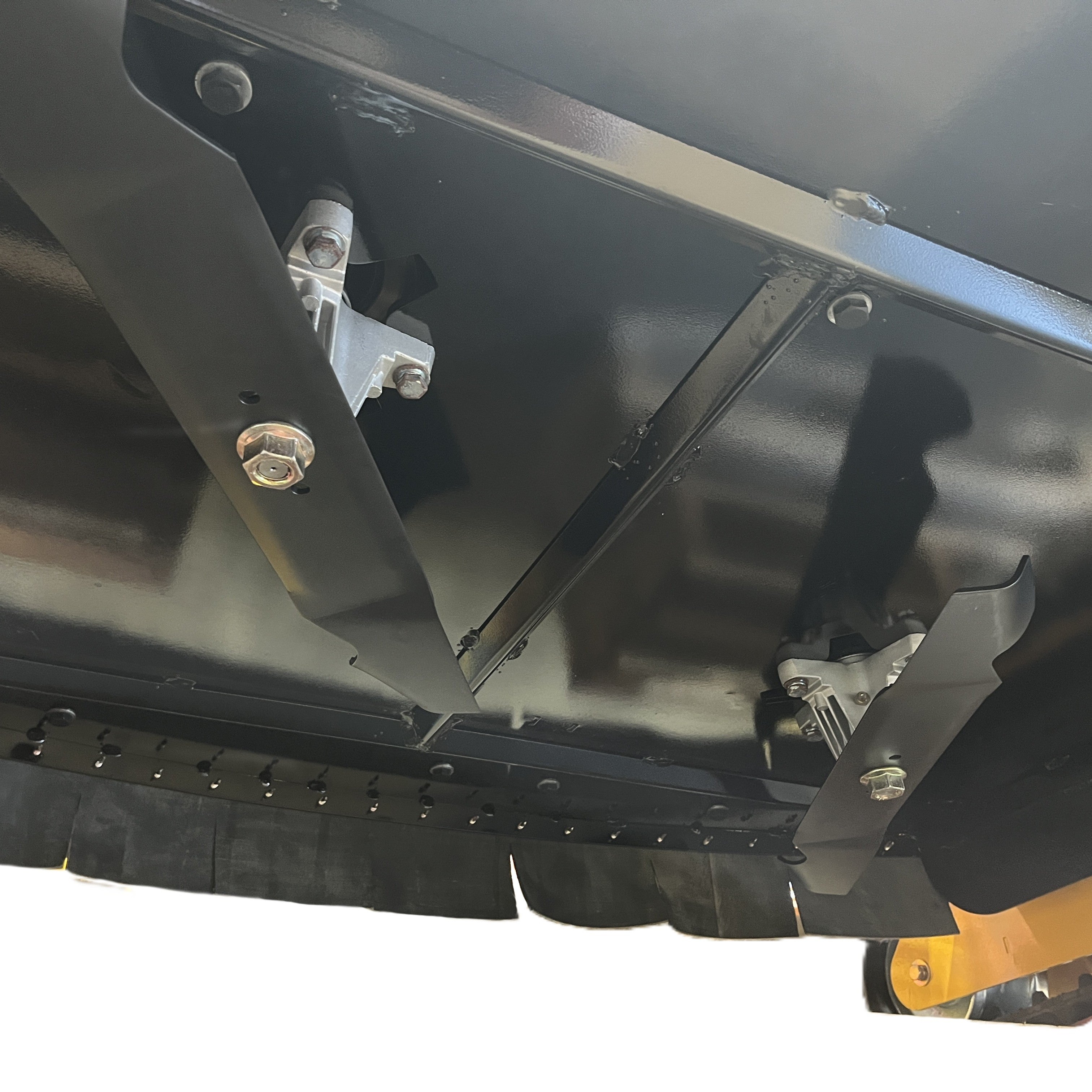 Close-up of the underside of a heavy machinery deck, showing two Paddock RC Mower Finishing Blade Set blades by Paddock mounted with bolts, assorted metal parts, and part of a protective rubber skirt along the edge.