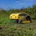 A yellow Paddock Remote Controlled Mower 7.5HP - 530mm with a track drive and 4-stroke petrol engine cuts tall grass in an outdoor field under a blue sky.