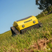 A Paddock Remote Controlled Mower 7.5HP - 530mm, in yellow, drives over green grass beneath a blue sky with trees in the background.