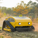 The Paddock Remote Controlled Mower 7.5HP - 530mm, by Paddock, operates autonomously across dry grass outdoors, with trees and shrubs in the background under natural light.