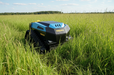 A Paddock Remote Controlled Mower 3kW Full Electric - 585mm in blue and black effortlessly cuts tall grass in a large open field beneath a blue sky with scattered clouds.