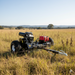 The Paddock Tow Behind Flail Mower by Paddock, featuring large wheels and red storage boxes, stands in a dry grassy field under a clear sky, powered by a Briggs and Stratton engine with hills and trees in the background.