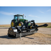 A green tractor is parked on a dirt road, while the Paddock Tractor PTO Flail Mower by Paddock, featuring chains and rotating parts, stands out in the foreground beneath a clear blue sky.