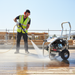 A construction worker in a safety vest uses the Paddock 3000 PSI Cold Water Pressure Washer by Paddock to clean a wooden floor at a building site, with construction materials and scaffolding visible in the background.