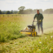 A person in a green shirt and gloves operates the Paddock Walk Behind Flail Mower by Paddock to cut tall grass for vegetation control in a sunny field bordered by trees and a wooden fence.