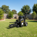 A Paddock Walk Behind Trencher with Guide Wheel by Paddock sits on a neatly mowed lawn in a fenced backyard, surrounded by trees and shrubs on a sunny day.