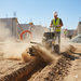 A construction worker in a hard hat and safety vest uses a Paddock Walk Behind Trencher with Guide Wheel to dig a narrow trench at a dusty construction site, with building frames and materials visible in the background.