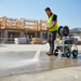 Wearing a safety vest and ear protection, a construction worker uses the Paddock 4,000 PSI Cold Water Pressure Washer by Paddock to clean concrete at a building site with wooden structures in the background.
