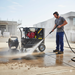 A man wearing ear protection uses a Paddock Hot Water Pressure Washer by Paddock to clean outdoor concrete at a construction site, with modern buildings and construction materials in the background.