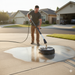A man uses the Paddock Driveway Cleaner by Paddock to wash a concrete driveway in a suburban neighborhood on a sunny day.