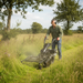 A man wearing ear protection uses a Paddock Walk Behind Tractor Flail Mower Attachment by Paddock to cut tall grass in a rural field surrounded by trees under a partly cloudy sky.