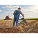 A farmer in blue overalls uses the Paddock Walk Behind Tractor Rotary Plough Attachment by Paddock to break up compacted soil in a large field, with a red barn and green crops visible under a partly cloudy sky.