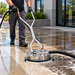 Wearing gloves and dark pants, an individual uses the Paddock™ Venturi Water Recovery Surface Cleaner by Paddock to wash an outdoor concrete area near a building, delivering efficient commercial pressure washing and revealing a distinct contrast between the cleaned and uncleaned sections.