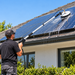 A person dressed in black is using the Paddock™ Rotating Brush Telescoping 5.5m Wand Kit – Solar Panel Cleaner by Paddock on a house roof to clean solar panels under a clear blue sky. Water runs off the panels as green shrubs appear in the foreground, showcasing the importance of maintaining elevated surfaces.