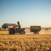 A rider in a helmet drives a red ATV towing the Paddock ATV & Quad Bike Trailer by Paddock, loaded with hay across a golden field, with a barn and hay bales in the background under a clear blue sky.