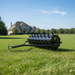The Paddock Lawn Spike Aerator by Paddock features spiked wheels, a towing handle, and a yellow "P" logo. It is pictured on a green lawn in front of a large stone house on a sunny day.