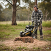 Wearing safety gear, a man operates a Paddock Commercial Stump Grinder by Paddock to remove an outdoor tree stump, surrounded by grass, trees, and scattered wood chips.