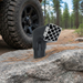 A Scintex Sherpa 4x4 Air Ram Snorkel Head with a silver grille is mounted on a black off-road vehicle parked on a pine needle-covered rock, with rugged tires and a forest trail in the background.