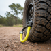 A close-up of a mud-covered off-road vehicle tire on gravel, featuring a Sherpa 4x4 Soft Rope Shackle looped through the wheel. Blurred trees and sky from the background are visible.