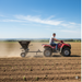 A man drives a red ATV across a plowed field on a sunny day, towing the Paddock Seed & Fertiliser Spreader (Heavy Duty) by Paddock. Young crops emerge from the soil as dust rises in the clear blue sky.