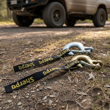 A pair of Sherpa 4x4 Winch Hooks with black and yellow straps are on the ground in front of an off-road vehicle parked on dirt, surrounded by forest.