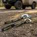 A pair of Sherpa 4x4 Winch Hooks with black and yellow straps are on the ground in front of an off-road vehicle parked on dirt, surrounded by forest.