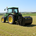 A green tractor with large yellow wheels is parked on a grassy field, attached to a Paddock Paddock Slasher with Catcher. Rolling hills and trees can be seen in the background under a clear sky.