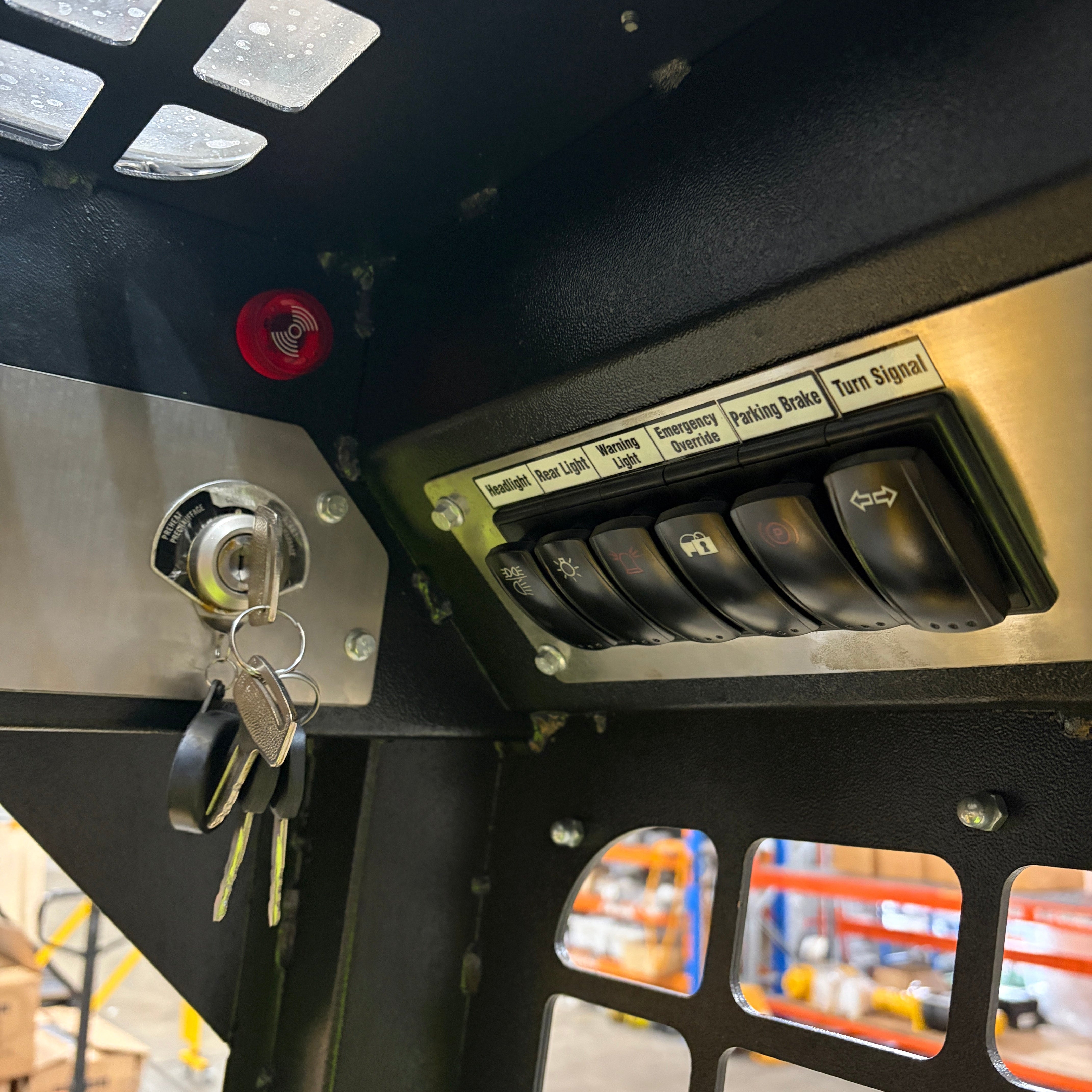Close-up of the Paddock Tracked Cabin Loader Pro control panel, showing keys in the ignition and labeled switches for headlights, hazard lights, parking brake, and turn signal. Metal grate and warehouse shelving are visible in the background.