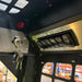 Close-up of the Paddock Tracked Cabin Loader Pro control panel, showing keys in the ignition and labeled switches for headlights, hazard lights, parking brake, and turn signal. Metal grate and warehouse shelving are visible in the background.