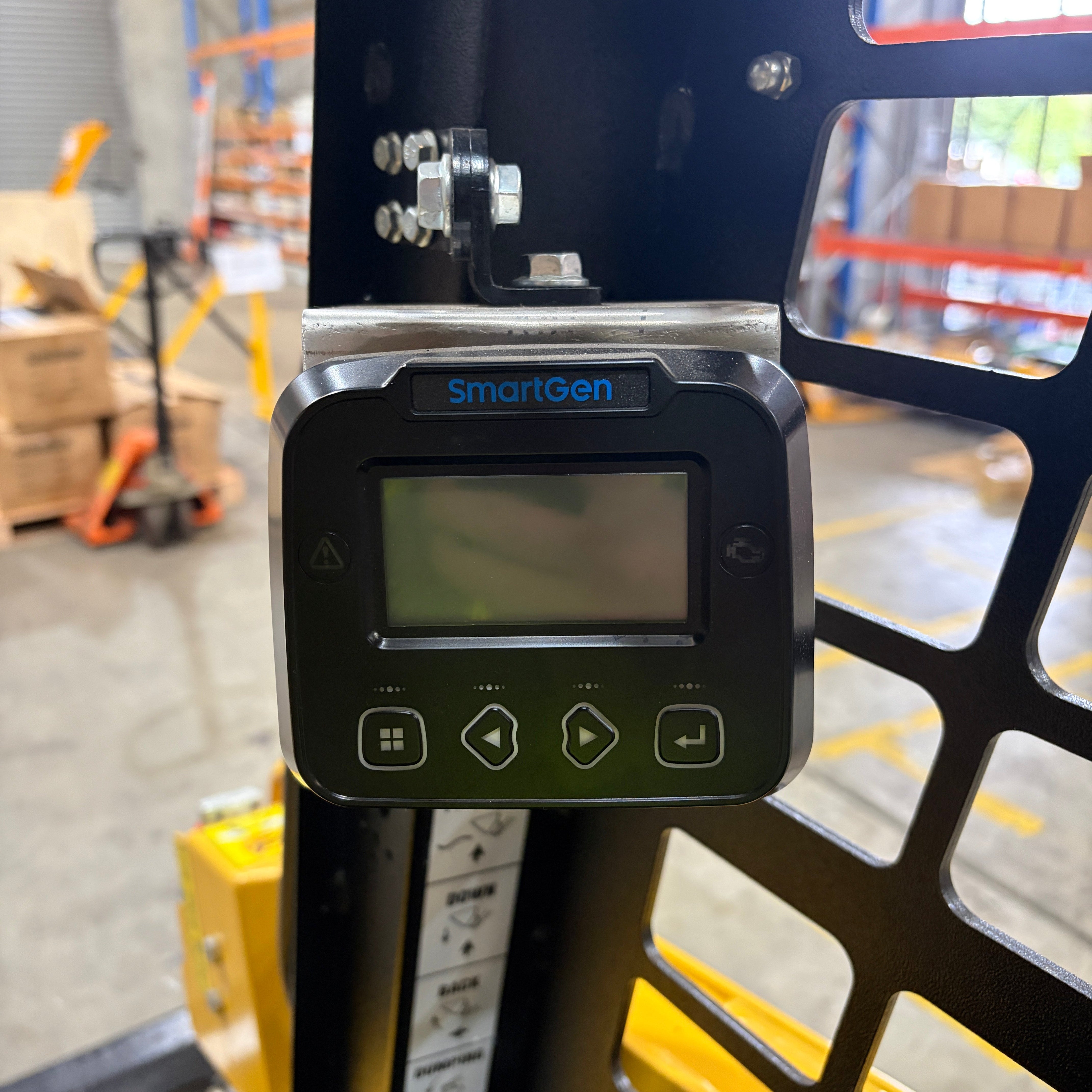 A close-up of a Paddock SmartGen digital control panel with screen and four buttons, mounted on a Paddock Tracked Cabin Loader Pro featuring a 40hp Perkins turbo diesel engine, in an industrial warehouse with shelves and boxes.