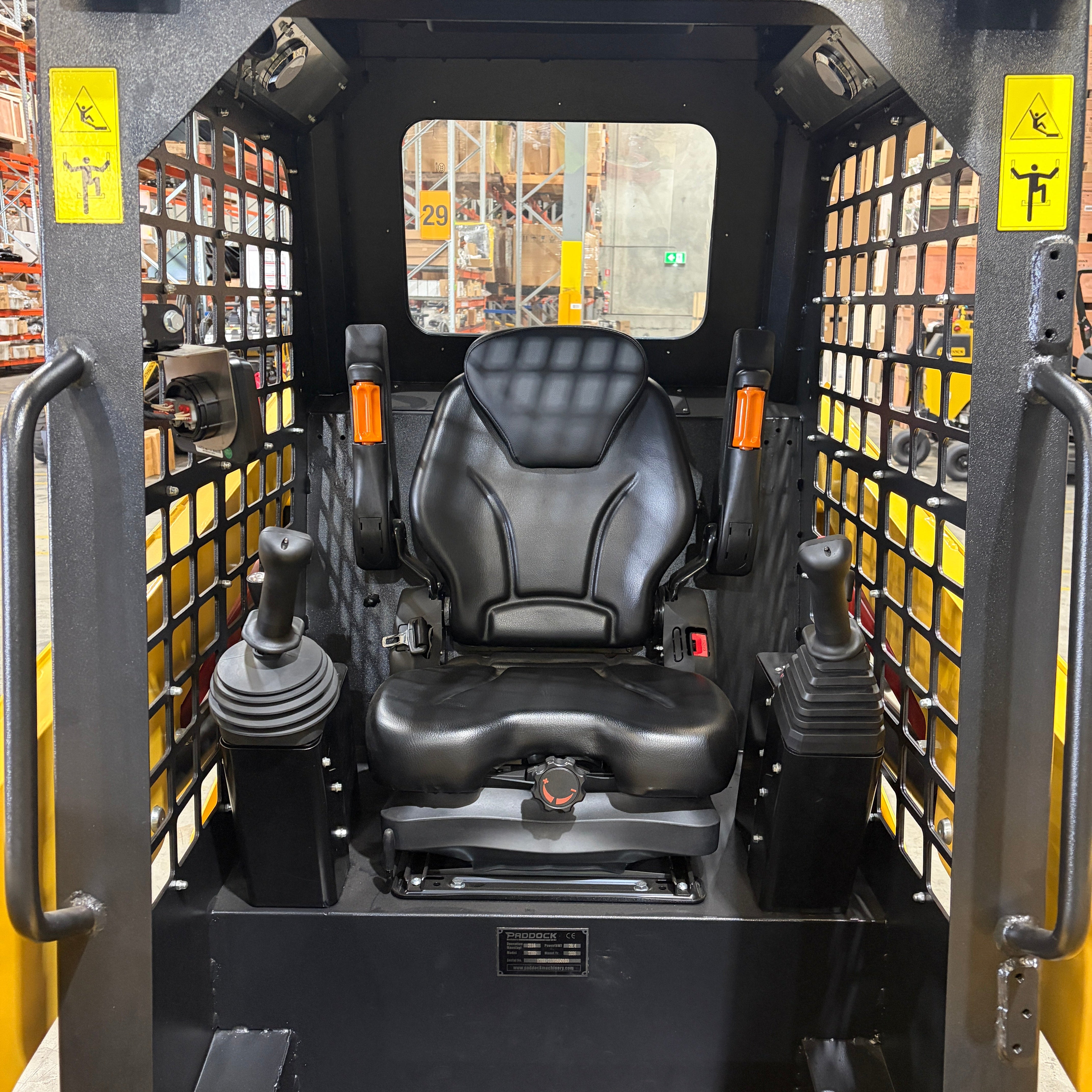 The image shows the operator seat and control joysticks inside a Paddock Tracked Cabin Loader Pro (40hp Perkins Turbo Diesel Engine, Ex Demo), with mesh side panels. Construction equipment shelves appear in the background.