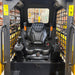 The image shows the operator seat and control joysticks inside a Paddock Tracked Cabin Loader Pro (40hp Perkins Turbo Diesel Engine, Ex Demo), with mesh side panels. Construction equipment shelves appear in the background.