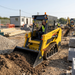 A construction worker operates a Paddock Tracked Cabin Loader Pro with a 40hp Perkins turbo diesel engine to move soil on a site. Building materials, a portable office, another worker, and an excavator are also visible among the equipment.