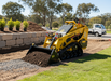 A Paddock Tracked Mini Loader Pro (40hp Perkins Turbo Diesel) transports soil in its bucket at a construction site, with stone landscaping, green grass, and trees in the background.