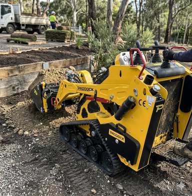 A Paddock Tracked Mini Loader Pro with a 40hp Perkins Turbo Diesel Engine moves soil at an outdoor landscaping site, demonstrating strong construction capability as workers and a truck can be seen in the background among trees and garden beds.