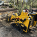 A Paddock Tracked Mini Loader Pro with a 40hp Perkins Turbo Diesel Engine moves soil at an outdoor landscaping site, demonstrating strong construction capability as workers and a truck can be seen in the background among trees and garden beds.