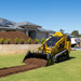 The Paddock Tracked Mini Loader (35hp Kubota Diesel) with Free 4in1 Bucket spreads soil on a lawn in front of a modern home featuring solar panels, stone retaining wall, potted plants, and a mulch pile beneath a clear blue sky.