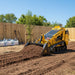 A Paddock Tracked Mini Loader (35hp Kubota Diesel Engine) with free 4in1 bucket spreads soil in a backyard near a wooden fence, shovels and large white material bags visible in the sunny background.