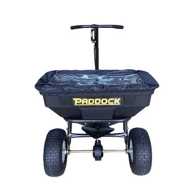 Front view of the Paddock Fertiliser & Seed Spreader, a black heavy-duty walk-behind unit with a handle, clear plastic cover, large rugged tires, and bold yellow Paddock branding, ideal for lawns, isolated on a white background.