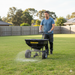 A man in gloves and a blue shirt uses the Paddock Fertiliser & Seed Spreader by Paddock to distribute material across a green suburban lawn bordered by wooden fencing.