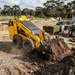 A Paddock Wheeled Mini Loader (35hp Kubota Diesel Engine) with Free 4in1 Bucket Bundle moves soil on a construction site, with dirt, gravel, stacked supplies, and trees in the background.